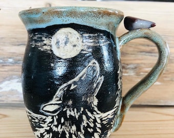 May include: A ceramic mug with a black background featuring a howling wolf under a full moon. The mug has a light blue rim and a decorative bird on the handle. The mug is on a wooden surface.