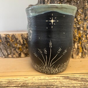 May include: A handmade ceramic mug with a black exterior and a light blue rim. The mug features a white star and wheat design. The mug is sitting on a wooden surface.