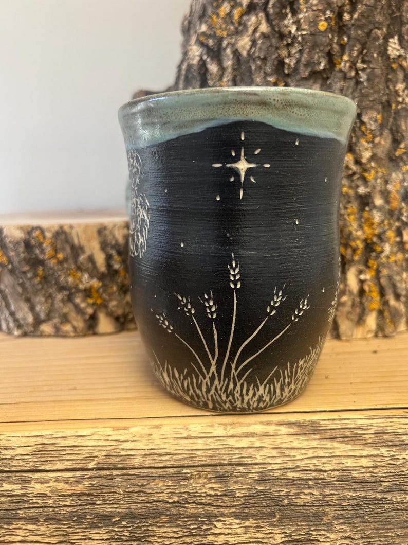 May include: A handmade ceramic mug with a black exterior and a light blue rim. The mug features a white star and wheat design. The mug is sitting on a wooden surface.