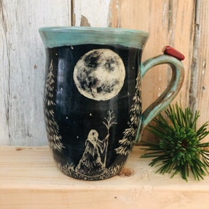 May include: Ceramic mug with a dark blue background featuring a full moon and a silhouetted figure holding a staff, surrounded by trees. The rim is teal, and a red stone adorns the handle. A unique, handcrafted piece.