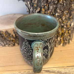 May include: A ceramic mug with a teal interior and handle. The exterior is black with a white tree and forest design. The mug sits on a wooden surface.