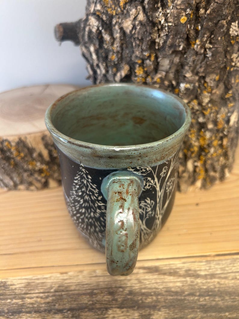 May include: A ceramic mug with a teal interior and handle. The exterior is black with a white tree and forest design. The mug sits on a wooden surface.