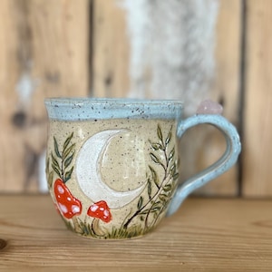 Peut inclure: Une tasse en céramique beige avec un bord et une anse bleu clair. La tasse présente un croissant de lune, des champignons rouges et blancs et du feuillage vert. Un cristal rose est fixé à la poignée.