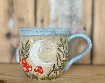 Może przedstawiać: Beżowy ceramiczny kubek z jasnoniebieską obręczą i uchwytem. Kubek przedstawia półksiężyc, czerwone i białe grzyby oraz zielone liście. Różowy kryształ jest przymocowany do uchwytu.