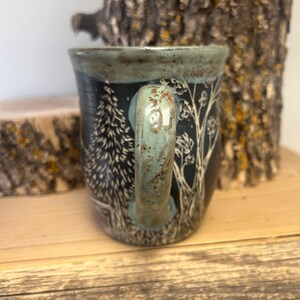 May include: A ceramic mug with a black background and a forest scene design. The handle and rim are a teal color. The forest scene includes trees and other foliage. The mug is sitting on a wooden surface.