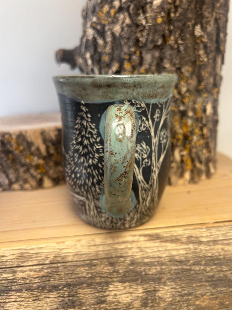 May include: A ceramic mug with a black background and a forest scene design. The handle and rim are a teal color. The forest scene includes trees and other foliage. The mug is sitting on a wooden surface.