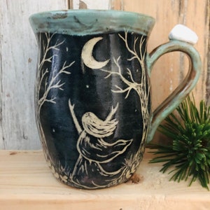 May include: A ceramic mug with a dark blue background featuring a white illustration of a woman with outstretched arms under a crescent moon and trees. The mug has a light blue rim and handle, with a white stone accent on the handle.