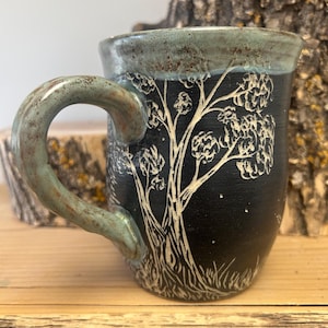 May include: A handmade ceramic mug with a black exterior and a tree design etched in white. The rim and handle have a teal glaze. The mug is sitting on a wooden surface.
