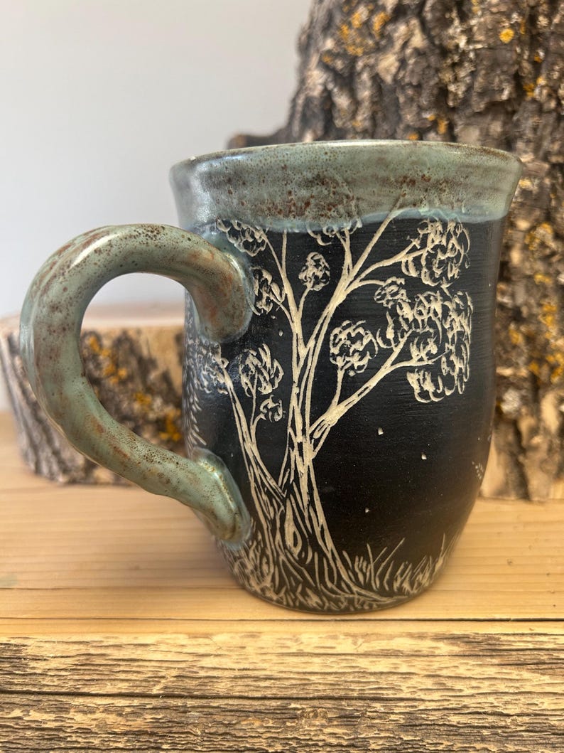 May include: A handmade ceramic mug with a black exterior and a tree design etched in white. The rim and handle have a teal glaze. The mug is sitting on a wooden surface.