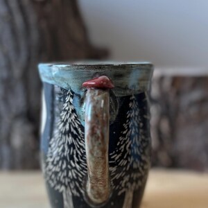 May include: A ceramic mug with a forest scene design. The mug is dark blue with white etched trees. The handle is brown with a red mushroom accent. The rim is a light blue.