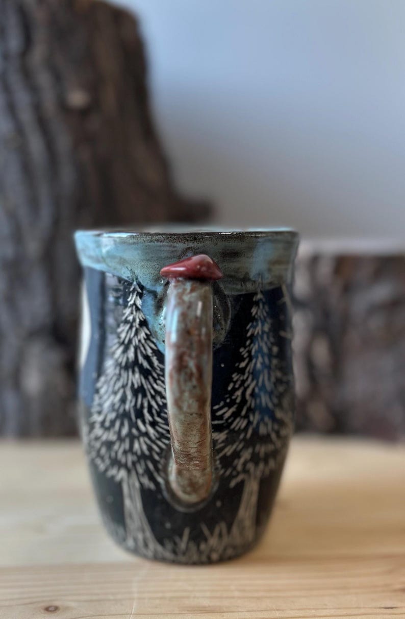 May include: A ceramic mug with a forest scene design. The mug is dark blue with white etched trees. The handle is brown with a red mushroom accent. The rim is a light blue.