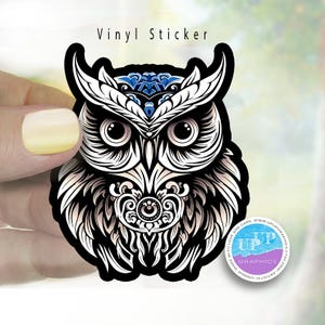 May include: A black and white vinyl sticker featuring a detailed illustration of an owl with blue accents. The owl's eyes are large and round, and its feathers are intricately patterned. The sticker has the text "Vinyl Sticker" at the top.