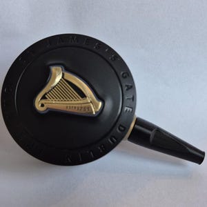 Op de afbeelding: Zwarte tapkraan met gouden harpembleem en de tekst "ST JAMES'S GATE DUBLIN". Het harpembleem heeft de tekst "ESTD 1759". De tapkraan is voor een drankje.