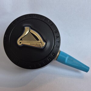 K&ouml;nnte beinhalten: Ein schwarzer Zapfhahn mit einem goldenen Harfenemblem und dem Text "ST JAMES'S GATE DUBLIN". Der Griff hat einen hellblauen Ausguss. Das Emblem ist auf einer kreisf&ouml;rmigen schwarzen Basis gepr&auml;gt.