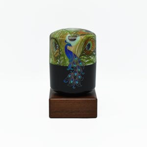 May include: A decorative black container with a peacock design. The top portion features a green and brown peacock feather pattern, while the bottom half is black with a blue peacock illustration. It sits on a small, square wooden base.
