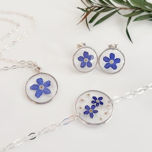 May include: A set of silver jewelry featuring forget-me-not flowers pressed inside clear resin. The set includes a necklace, a bracelet, and a pair of earrings.