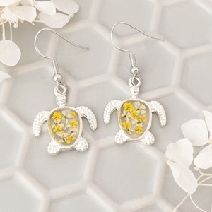 May include: A pair of silver dangle earrings featuring a turtle design with pressed yellow and white flowers inside the resin.