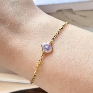 May include: A delicate gold-colored chain bracelet featuring a small, clear resin charm. The charm is shaped like a flower and contains a small, blue flower. The bracelet is worn on a wrist.