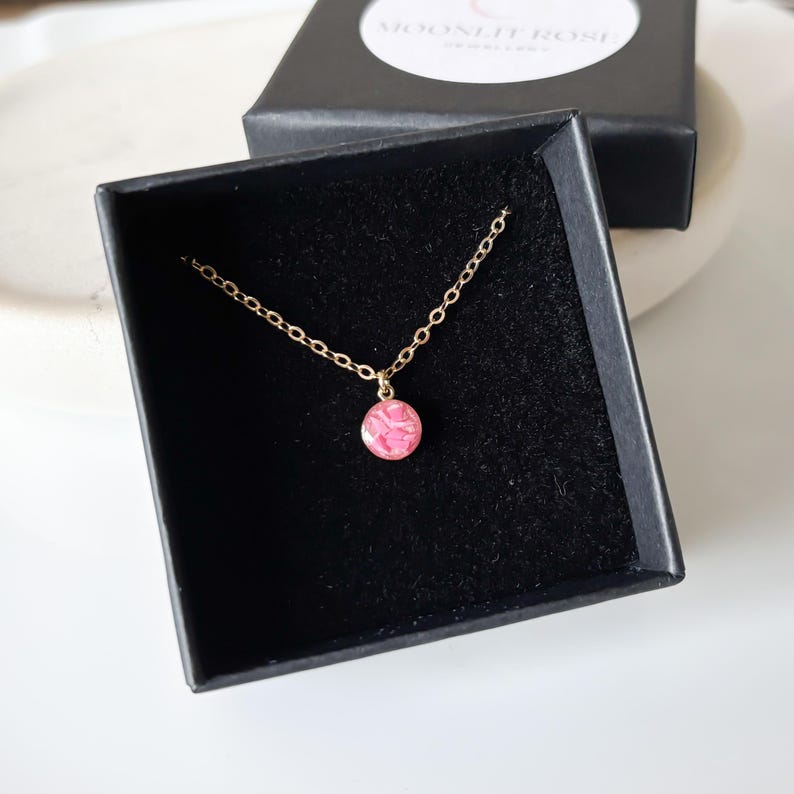 May include: A delicate gold chain necklace with a small pink circle pendant. The pendant has a swirling pattern inside.