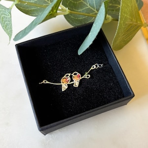 May include: A silver necklace featuring two small birds perched on a branch. The birds are filled with pressed flowers in shades of orange, red, and white.