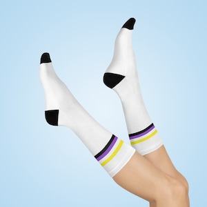 Peut inclure: Une paire de chaussettes blanches avec des talons noirs et une bande violette et jaune. Les chaussettes sont portées sur les jambes d'une personne.