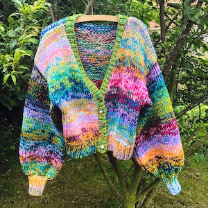 Puede incluir: Un cárdigan de punto colorido con cuello en V y cierre de botones. El cárdigan tiene un patrón de arcoíris con tonos de rosa, azul, verde, amarillo y morado.