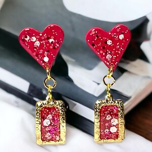 May include: A pair of red heart-shaped dangle earrings with gold accents. The hearts are covered in red glitter and the dangle is a gold rectangle with red glitter inside.