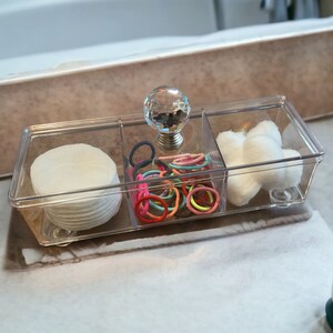 May include: A clear, rectangular organizer with three compartments. One section holds cotton pads, another contains colorful hair ties, and the third has cotton balls. The organizer features a crystal knob and is designed for bathroom or vanity use.