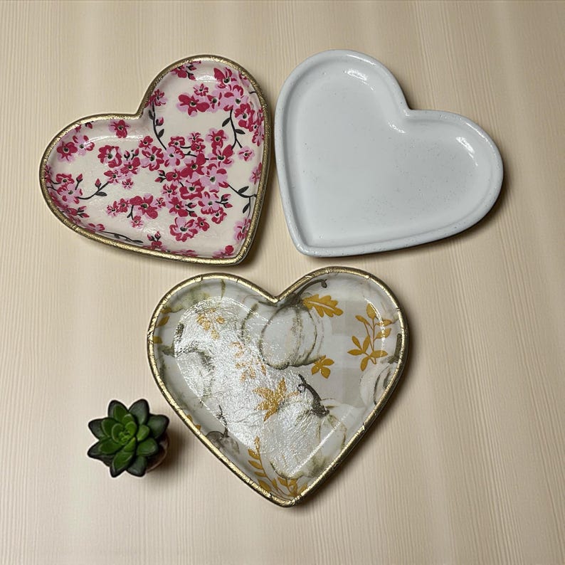 Pu&ograve; includere: Tre vassoi decorativi a forma di cuore. Un vassoio presenta un motivo floreale con fiori rosa e un bordo dorato. Un altro &egrave; bianco, e il terzo ha un design a zucca con accenti dorati e un bordo dorato. &Egrave; presente anche una piccola pianta succulenta.