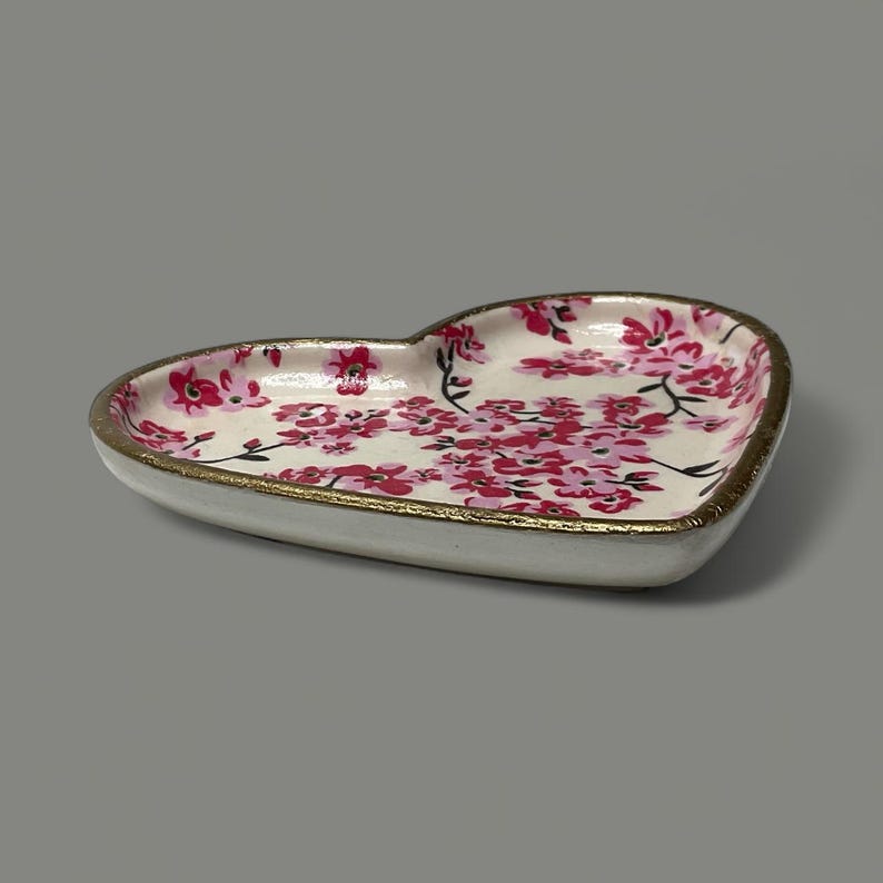 Pu&ograve; includere: Piatto in ceramica a forma di cuore con un motivo floreale nei toni del rosa e del bianco. Il bordo &egrave; rifinito con oro. L'oggetto &egrave; decorativo.