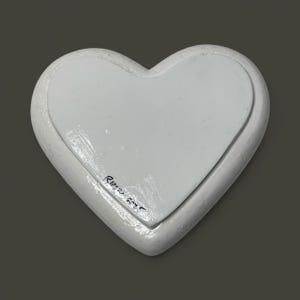 Pu&ograve; includere: Un oggetto decorativo bianco a forma di cuore con un bordo rialzato e un centro incassato. L'oggetto ha una finitura liscia e verniciata. La firma dell'artista e l'anno 2023 sono visibili nell'angolo in basso a sinistra. Lo sfondo &egrave; grigio.
