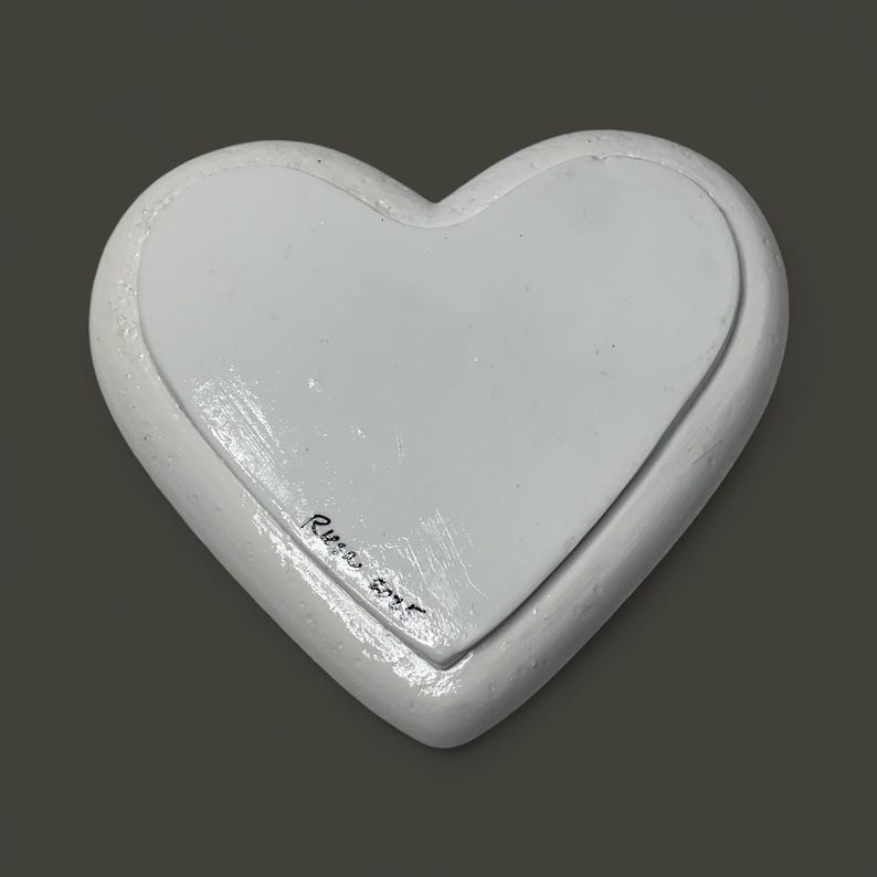 Pu&ograve; includere: Un oggetto decorativo bianco a forma di cuore con un bordo rialzato e un centro incassato. L'oggetto ha una finitura liscia e verniciata. La firma dell'artista e l'anno 2023 sono visibili nell'angolo in basso a sinistra. Lo sfondo &egrave; grigio.