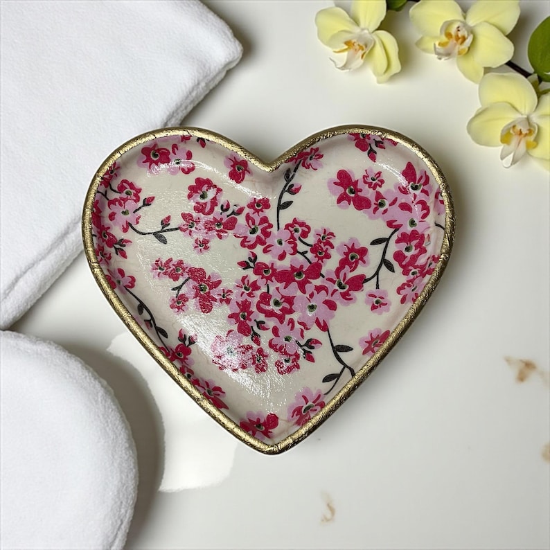 Pu&ograve; includere: Piatto decorativo a forma di cuore con un motivo floreale nei toni del rosa e del rosso su sfondo crema. Il piatto ha un bordo dorato. L'immagine mostra anche asciugamani bianchi e orchidee gialle.
