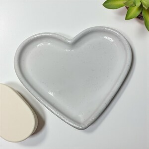 Pu&ograve; includere: Un piatto in ceramica a forma di cuore grigio chiaro macchiettato. Il piatto ha un bordo rialzato e un interno liscio. Un piccolo oggetto a forma di goccia, bianco sporco, &egrave; visibile a sinistra del piatto a forma di cuore. Una piccola pianta succulenta.