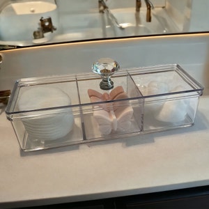 May include: A clear plastic organizer with three compartments. The organizer has a crystal-like knob on the top and contains cotton rounds, white cotton balls, and two pink butterfly-shaped soaps.