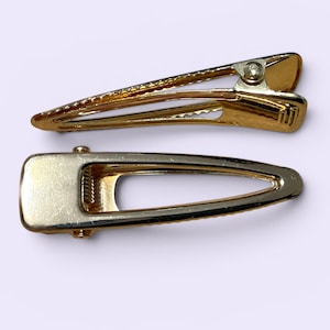 May include: Two gold-toned metal hair clips. The top clip is a long, slender triangular shape with a serrated edge. The bottom clip is a wider, elongated oval shape with a smooth, flat surface and a spring-loaded hinge mechanism at the base.