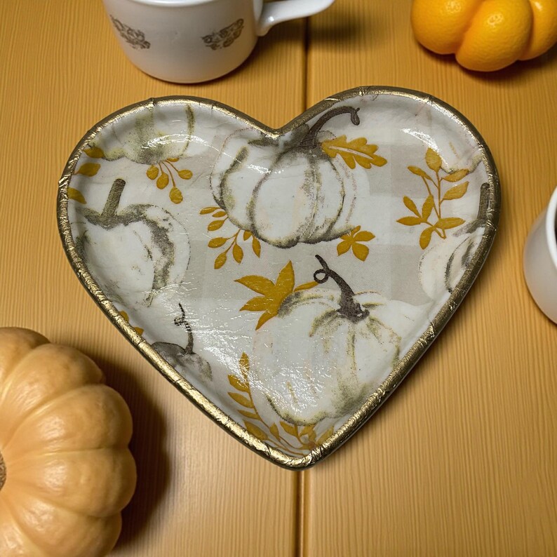 Pu&ograve; includere: Piatto decorativo a forma di cuore con bordo dorato, con un motivo di zucche bianche su sfondo a quadri grigio chiaro. Il piatto &egrave; circondato da zucche e tazze, creando un tema autunnale.
