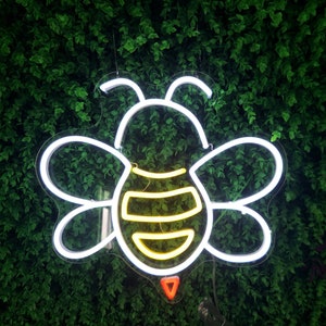 Puede incluir: Un contorno de abeja de ne&oacute;n blanco con un cuerpo de ne&oacute;n amarillo y naranja sobre un fondo de hojas verdes.