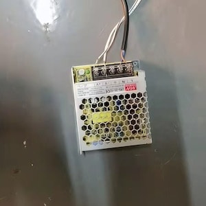 Könnte beinhalten: Ein silbernes Netzteil mit Wabenmuster-Belüftung. Mehrere Drähte, darunter schwarze, weiße und braune, sind angeschlossen. Das Gerät ist auf einer grauen Oberfläche montiert. Maße in cm.