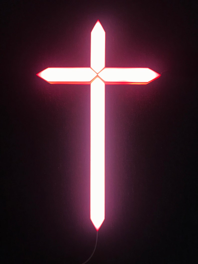 May include: A white cross-shaped light fixture with a pink glow, hanging from a black cord against a dark background.