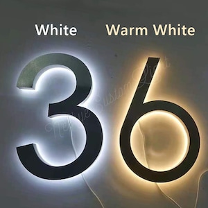 Puede incluir: Dos números de casa de metal negro, 3 y 6, con un contorno de luz LED blanca. El número 3 está iluminado con una luz blanca brillante, mientras que el número 6 está iluminado con una luz blanca cálida.