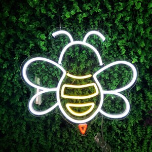 Puede incluir: Luz de ne&oacute;n blanca con forma de abeja con un cuerpo a rayas amarillo y blanco sobre un fondo de hojas verdes.
