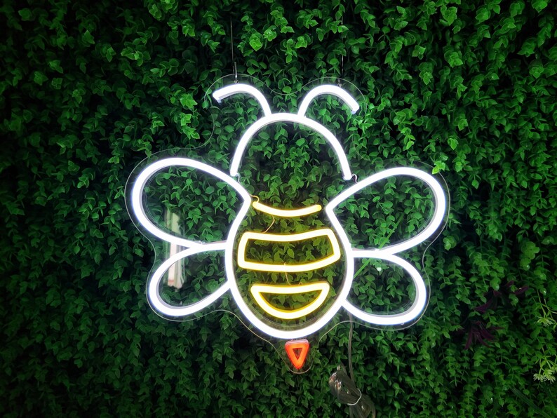 Puede incluir: Luz de ne&oacute;n blanca con forma de abeja con un cuerpo a rayas amarillo y blanco sobre un fondo de hojas verdes.