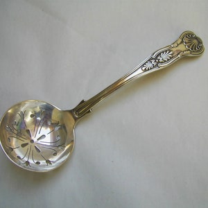 May include: A silver sugar spoon with an ornate handle and a perforated bowl. The bowl has a decorative design of a starburst with floral accents.