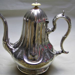 May include: A silver teapot with a fluted lid and an ornate handle. The teapot has a decorative design and a polished finish.