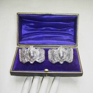 May include: Two silver napkin rings with ornate designs, featuring a heart-shaped centre, are displayed in a purple velvet-lined box.