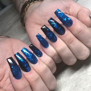 Peut inclure: Gros plan sur des ongles longs et brillants, noirs et bleus, sur la main d'une personne. Les ongles sont longs et ont une forme de cercueil. Le brillant est bleu et noir et est appliqué en dégradé.