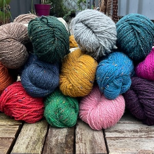 Può includere: Una collezione di 15 gomitoli di filato in vari colori, tra cui viola, arancione, rosso, verde, blu, grigio, rosa e marrone. Il filato è disposto a forma di piramide su una superficie di legno.