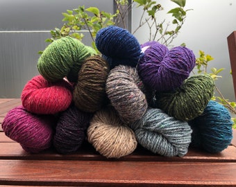 May include: A pile of colorful yarn skeins in various shades of green, blue, purple, red, brown, and gray. The yarn is arranged in a pyramid shape on a wooden surface.