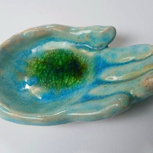 May include: A hand-shaped ceramic trinket dish featuring a glossy glaze in shades of light blue and turquoise. The center of the palm displays a vibrant, dark green crackle glaze finish, creating a distinct contrast against the lighter blue tones.